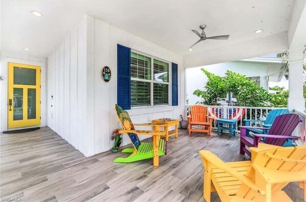 A covered outdoor seating area with colorful Adirondack chairs, a wooden table, and a ceiling fan.