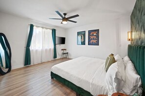 A hotel room with a bed, a ceiling fan, a TV, and artwork on the wall.