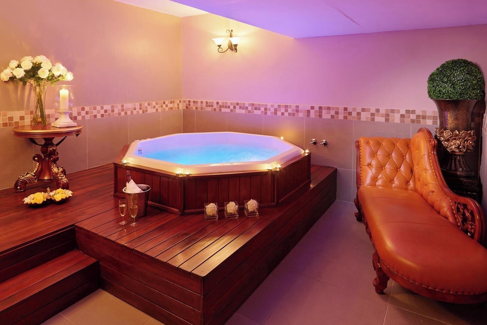 Een spa-ruimte met een houten terras, een whirlpool, een ligbed en een wandlamp.