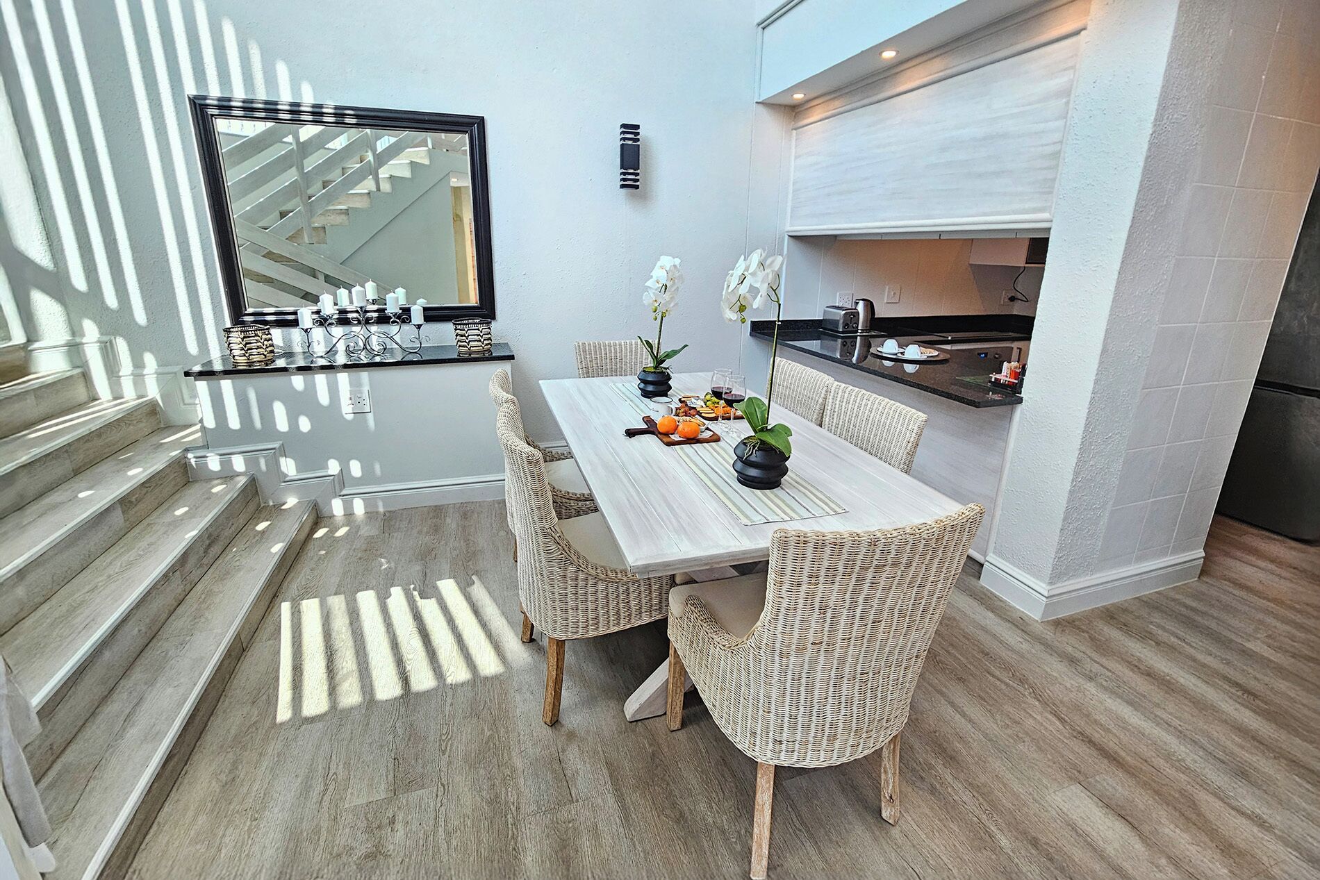 A modern kitchen with a dining area, featuring a light-colored wooden table and wicker chairs, a built-in stove, and a large window with a view of a staircase.