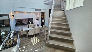 A modern interior with a staircase leading to an upper level, a dining area with a table set for two, and a kitchen with a large window. - Penthouse 711 at La Montagne by First Private Stays (Ballito)