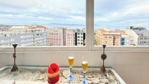 Un balcon avec une table où sont exposés des coquillages, un chapeau rouge et deux verres de bière. On aperçoit un paysage urbain en arrière-plan.