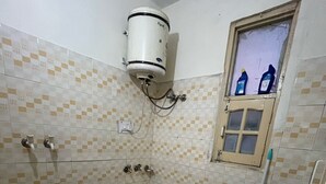 Une petite salle de bain avec des toilettes, un seau vert, une fenêtre avec des bouteilles et un chauffe-eau fixé au mur.