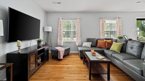 A modern living room with a flat-screen TV, a sofa, a coffee table, and two windows with curtains.