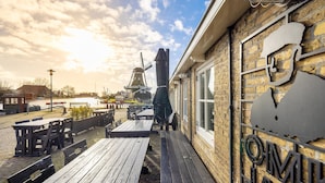 Une terrasse en bois avec des bancs et des tables, un moulin à vent en arrière-plan et un bâtiment à la façade en briques et doté d’une enseigne métallique.