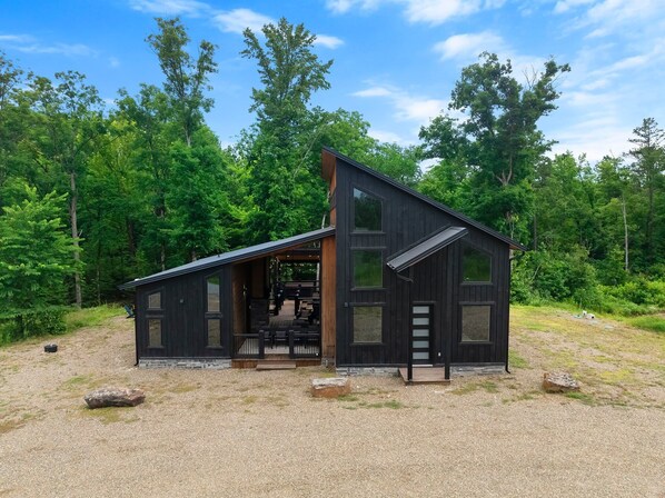 A modern cabin with a large porch, multiple windows, and a gravel driveway.