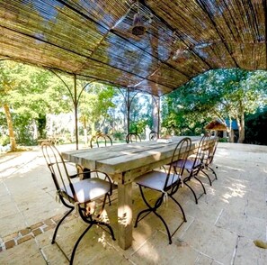 An outdoor patio with a wooden table and metal chairs, surrounded by stone flooring and shaded by a woven canopy.