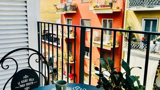 Un balcone con un tavolo apparecchiato per due, con dolci e tazzine di caffè, affacciato su un edificio colorato.
