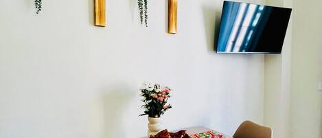 A dining area with a table set for two, chairs, a vase with flowers, and a flat-screen TV mounted on the wall.