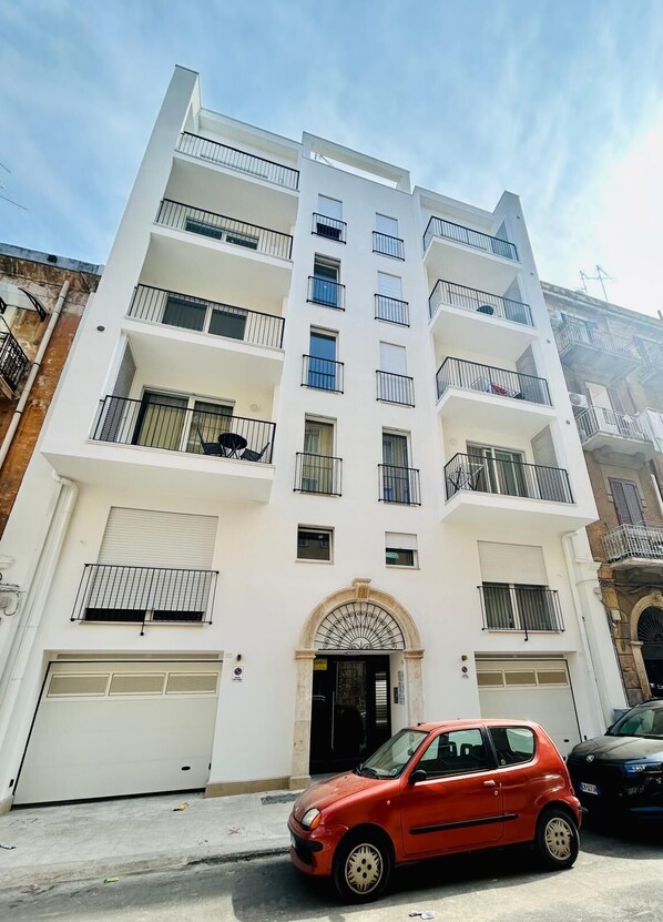 Un immeuble à plusieurs étages, aux murs extérieurs blancs, doté de nombreux balcons et d’une entrée centrale surmontée d’une arche.