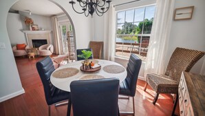 Un coin repas avec une table ronde, des chaises noires et un lustre. On y trouve également une cheminée, des fauteuils en osier et une vue sur l’extérieur grâce à de grandes fenêtres.