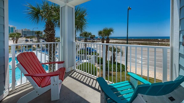 A balcony with red and blue Adirondack chairs, a pool, and a view of the beach and ocean.