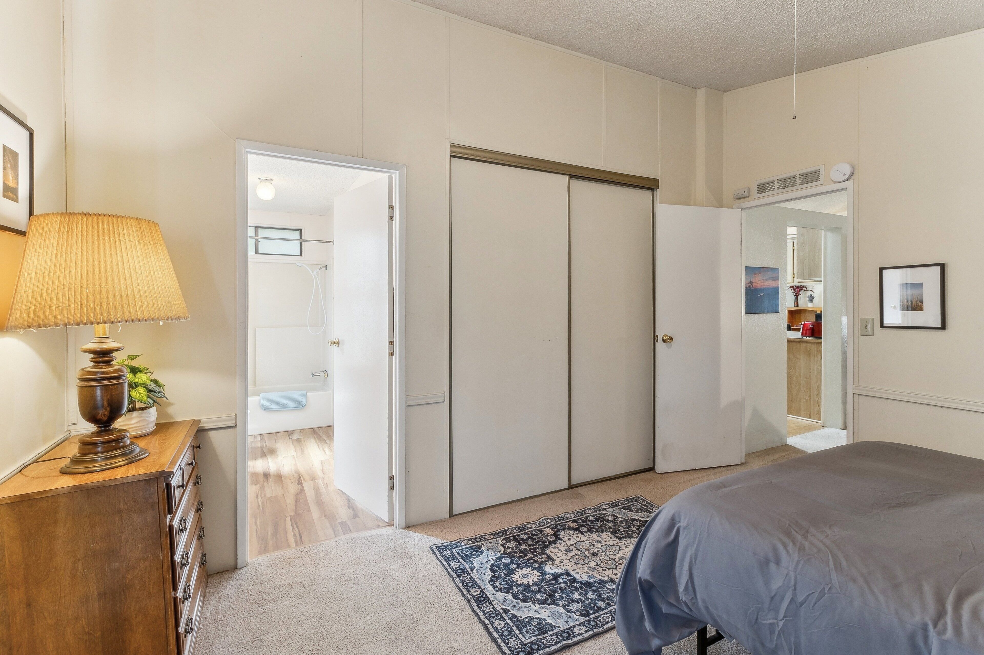 Une chambre à coucher comprenant un lit, une commode, une lampe, une porte donnant sur une salle de bain et un miroir au mur.