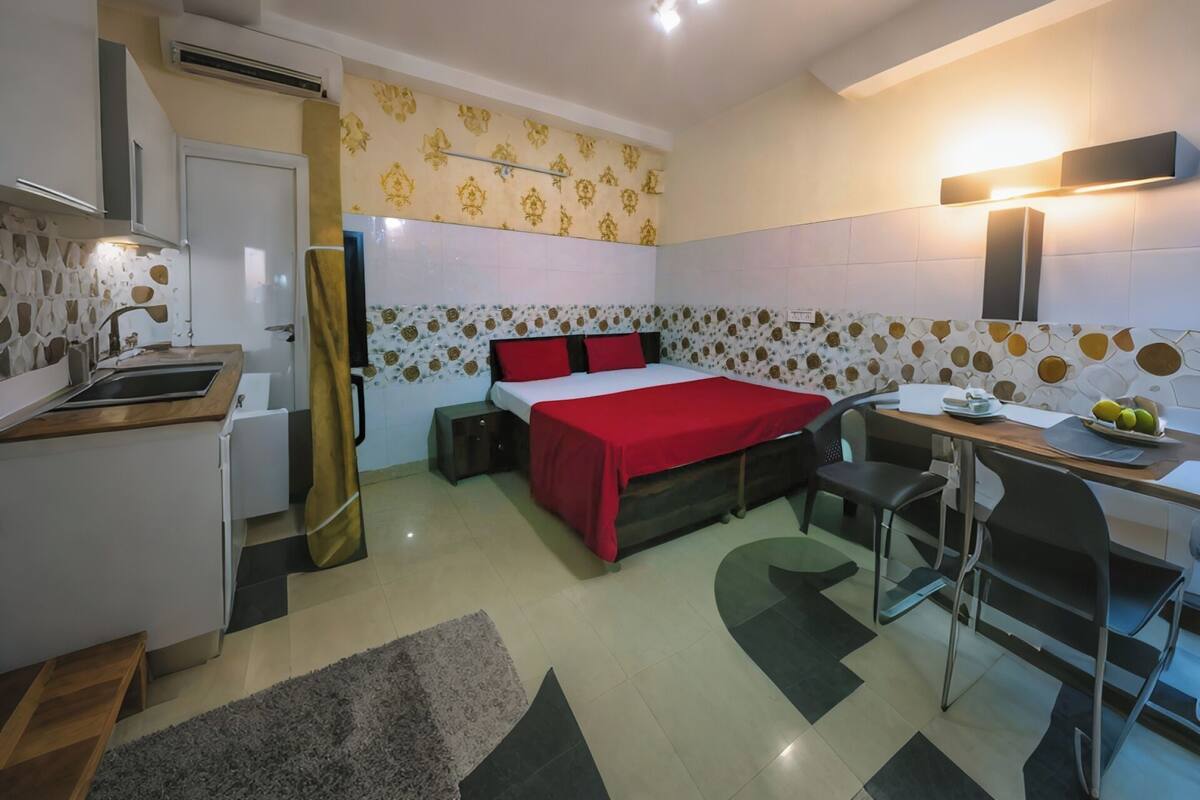 A compact hotel room with a bed, a desk, a chair, a small kitchenette, and a bathroom.