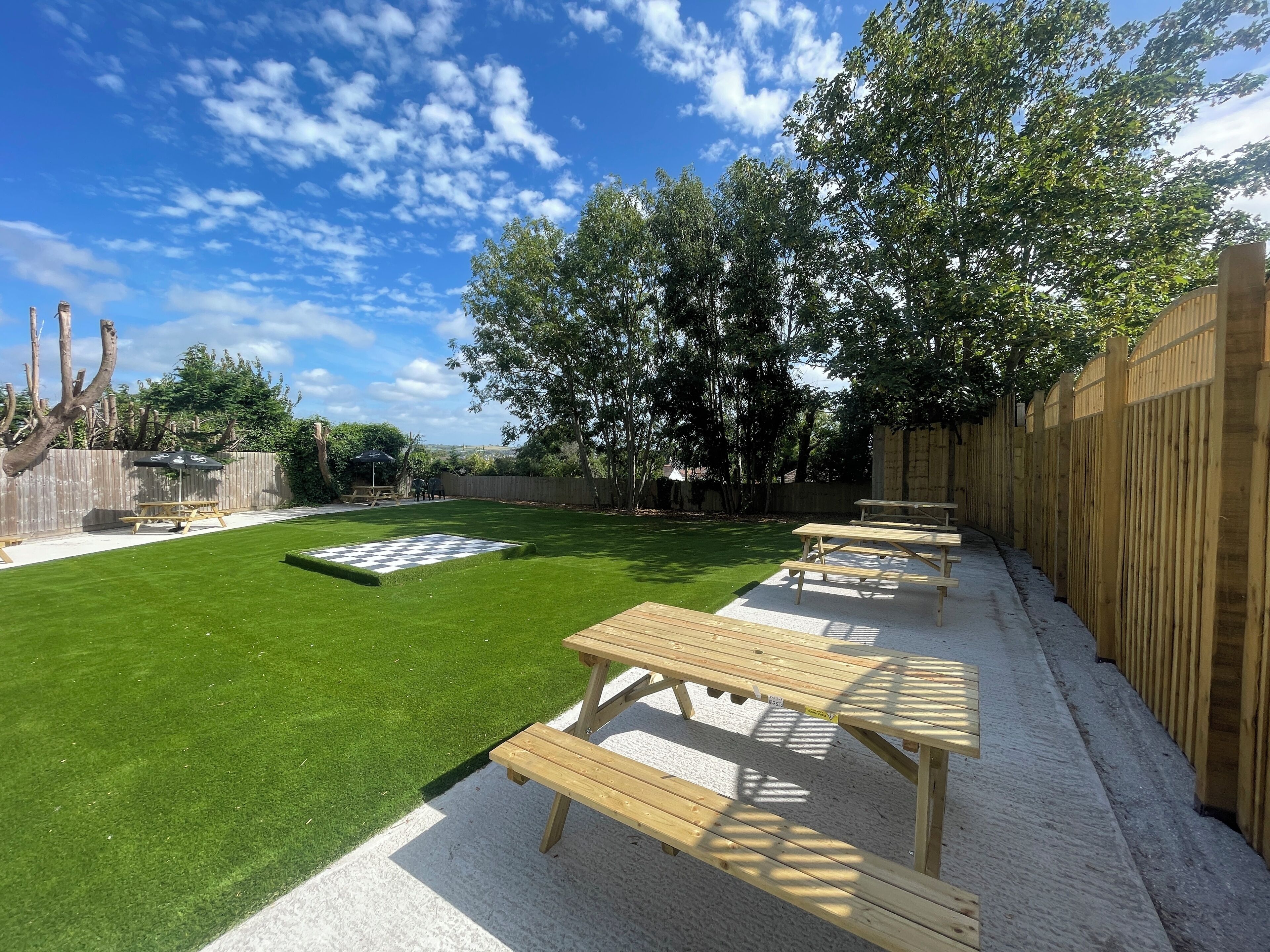 A backyard with a green lawn, a checkered area, and wooden picnic tables.