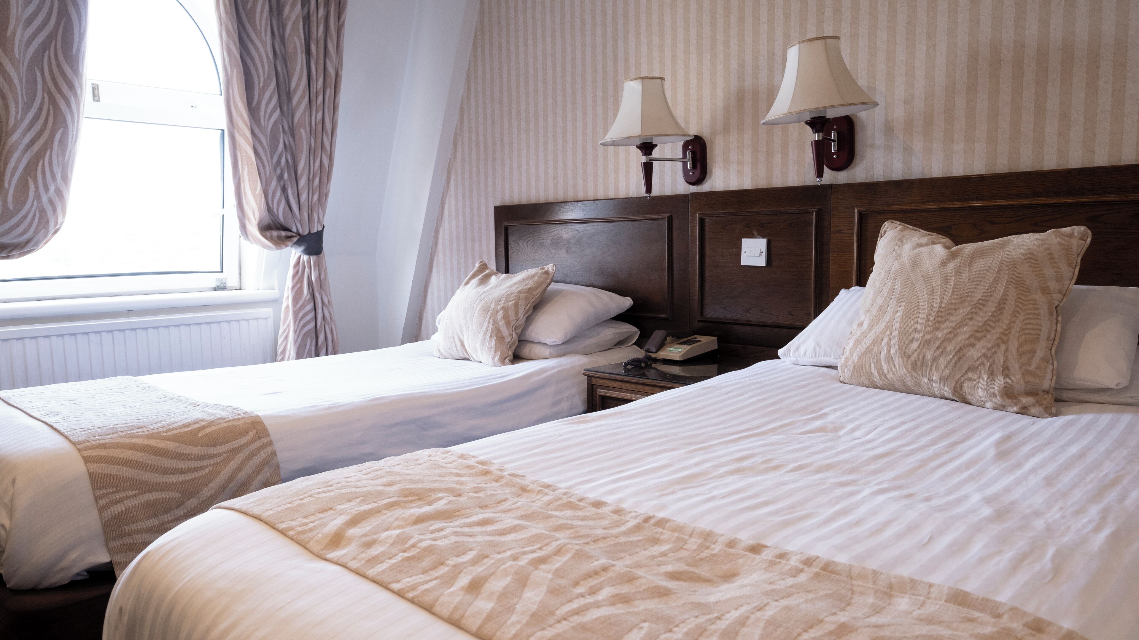 A hotel room with two beds, a nightstand with a lamp, and a window with curtains.