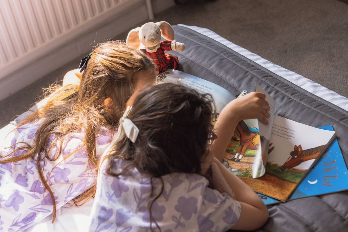 Two children reading a book on a bed with a pillow.