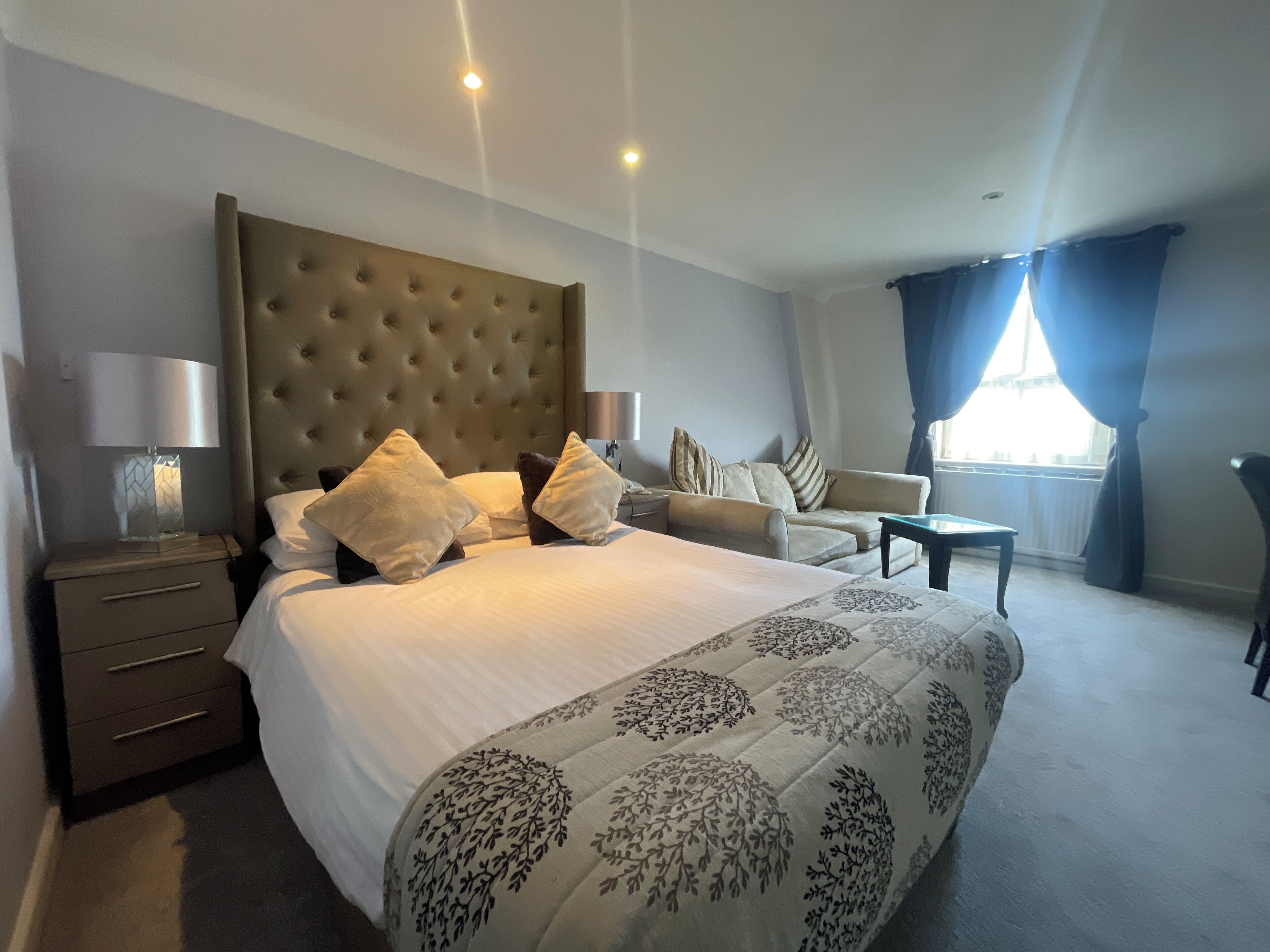 A hotel room with a large bed, a sofa, a small table, and a chair. There are two lamps on the nightstands.