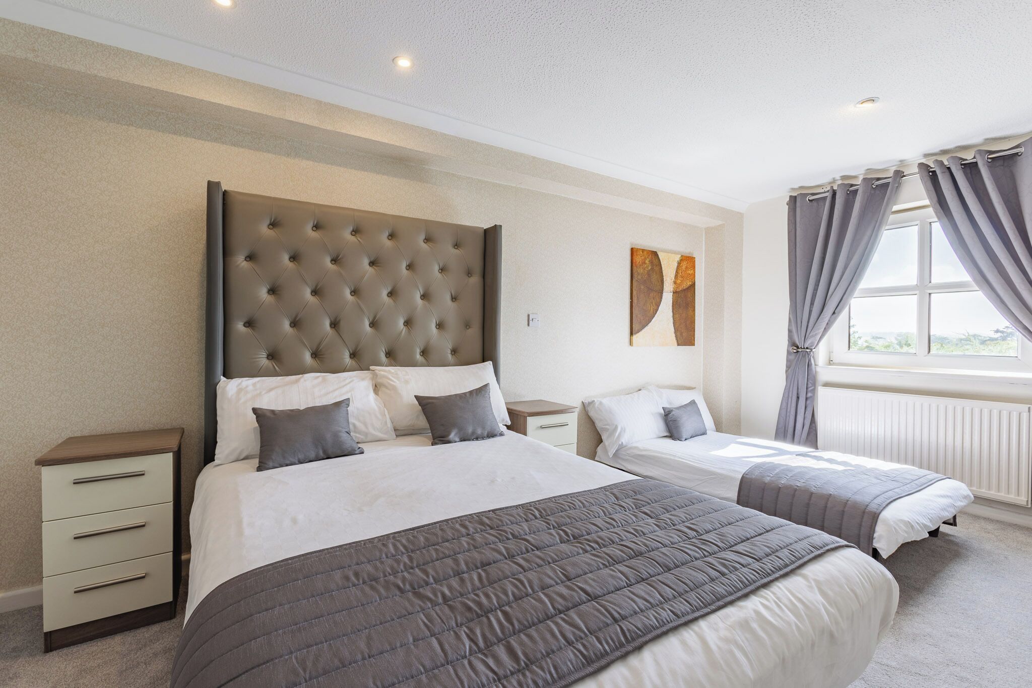 A hotel room with two beds, a large tufted headboard, a nightstand with drawers, and a window with curtains.