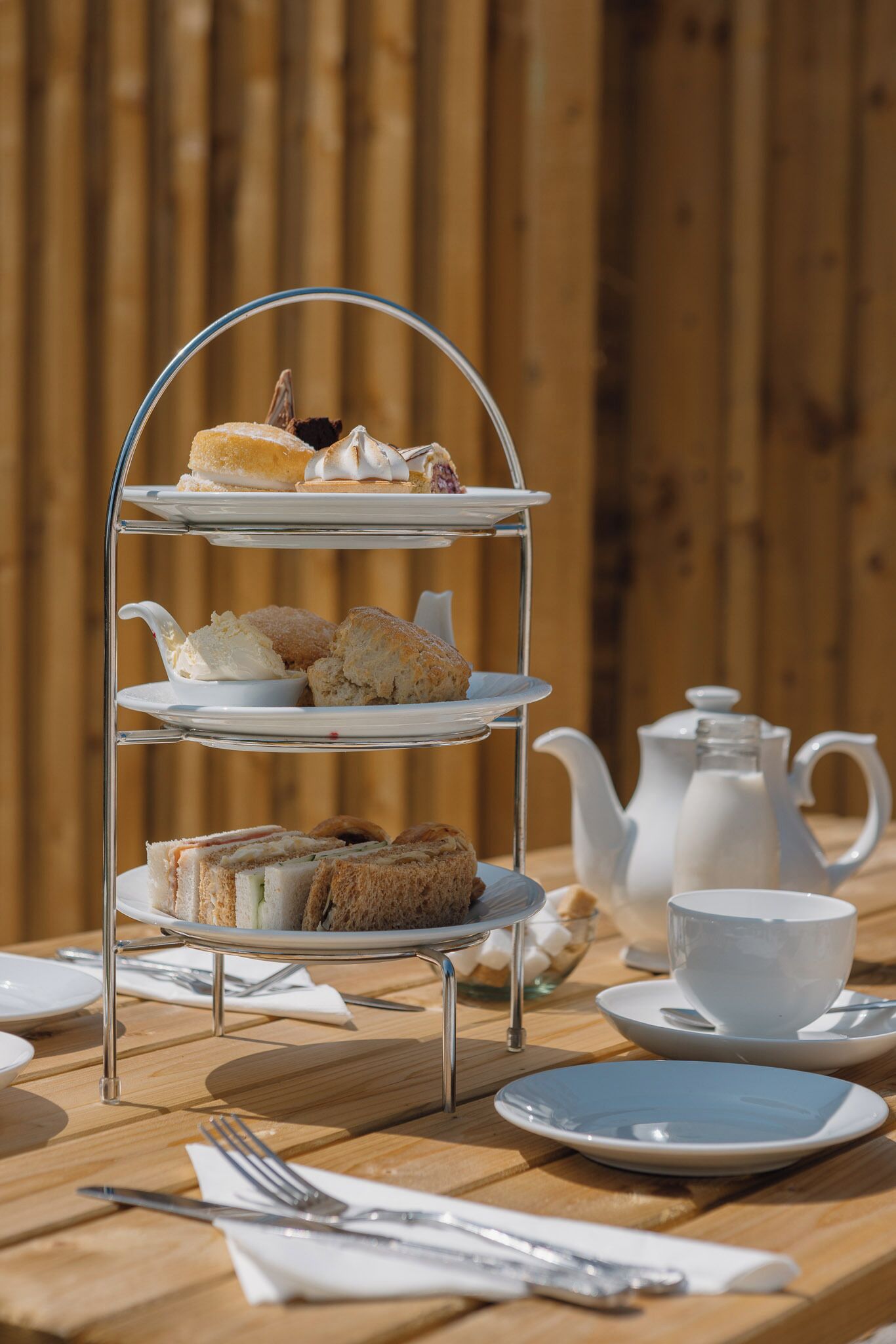 A three-tiered dessert stand with various pastries and sandwiches, a teapot, and cups on a wooden table.
