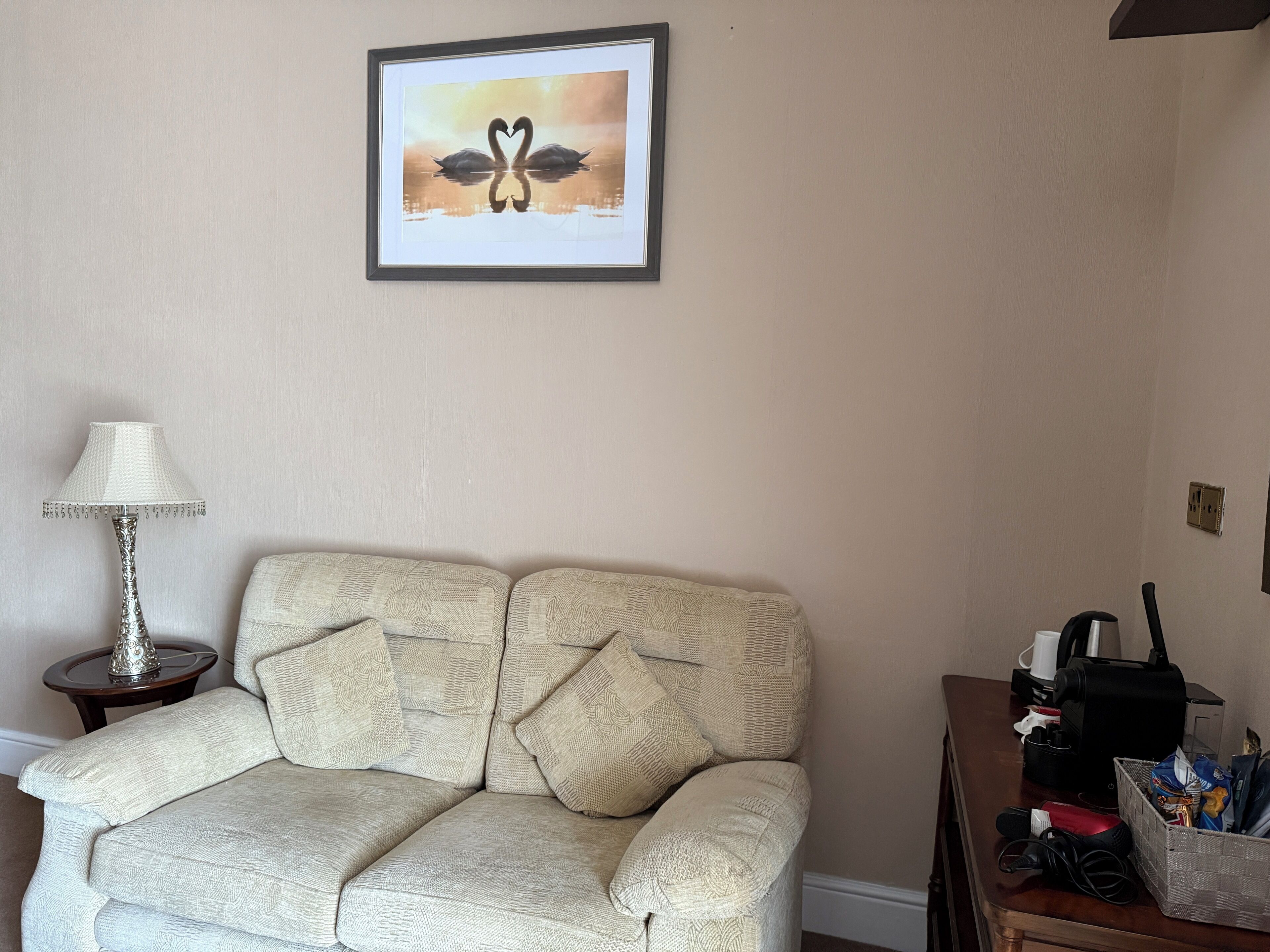 A beige sofa with a textured pattern, a framed picture of swans on the wall, a lamp on a side table, and a desk with various items including a coffee maker and a basket.