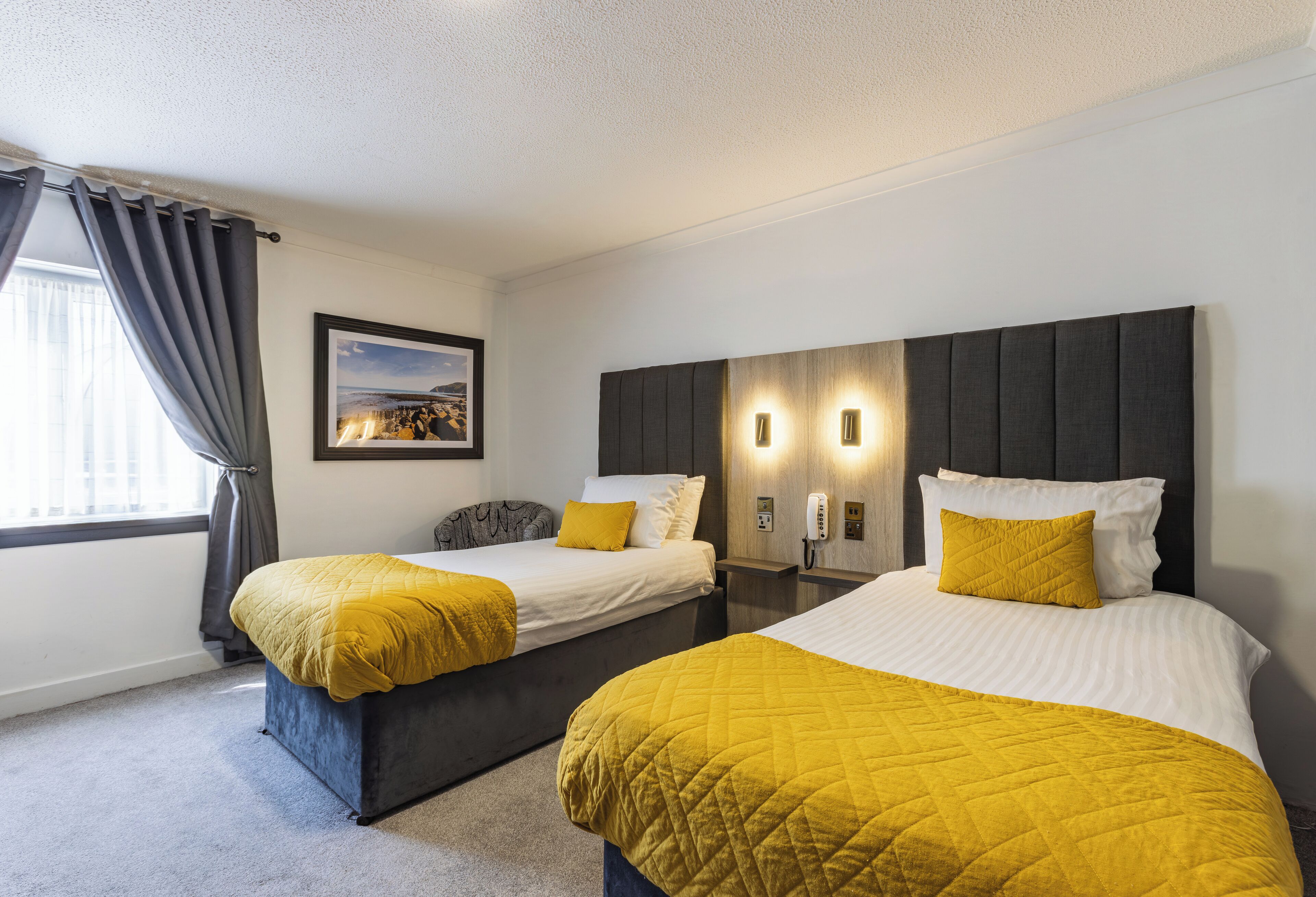 A hotel room with two beds, each with a yellow quilted bedspread and a white pillow. There is a framed picture on the wall, a bedside table with a lamp, and a window with curtains.