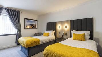 A hotel room with two beds, each with a yellow quilted bedspread and a white pillow. There is a framed picture on the wall, a bedside table with a lamp, and a window with curtains.