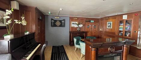 A room with a piano, a dartboard, a fireplace, and a seating area with a table and chairs.