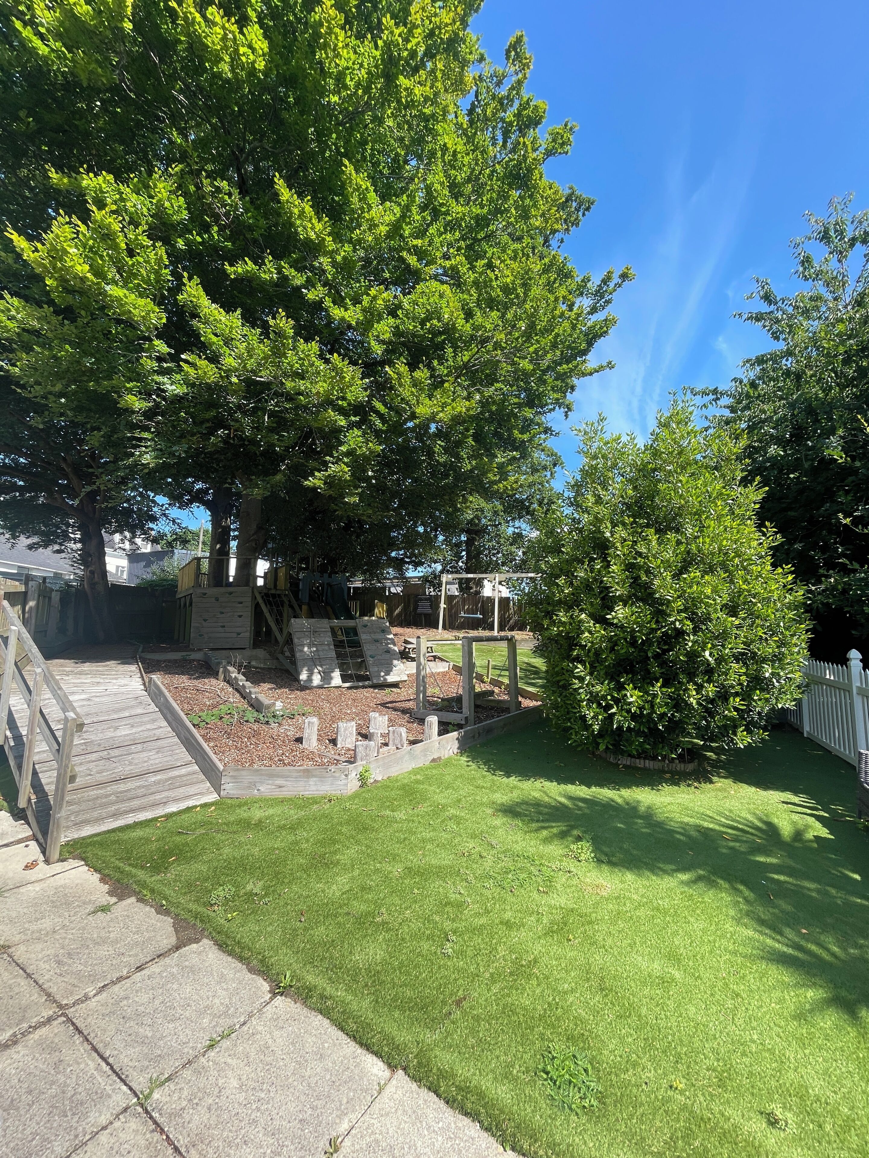 Un patio trasero con una terraza de madera, una pequeña jardinera y una cerca blanca de piquetes.