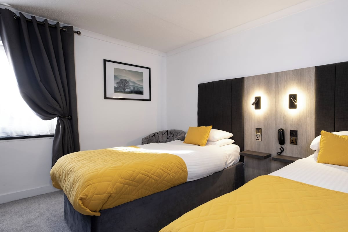 A hotel room with two beds, a large window with curtains, a framed picture on the wall, and a modern light fixture above the beds.