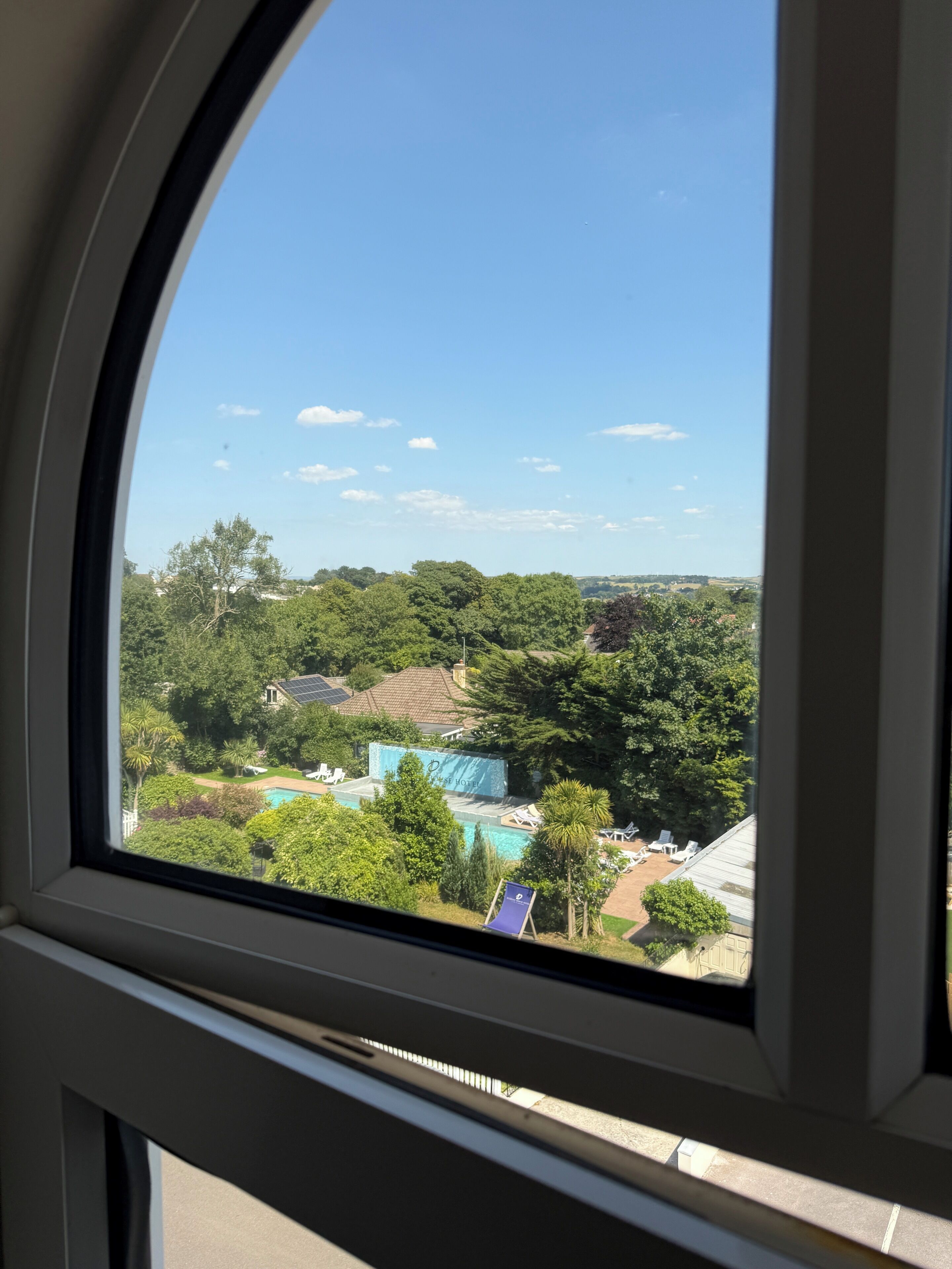 A view through a window of a property with a swimming pool, lush greenery, and a clear sky.
