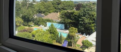 A view through a window of a property with a swimming pool, lush greenery, and a clear sky.