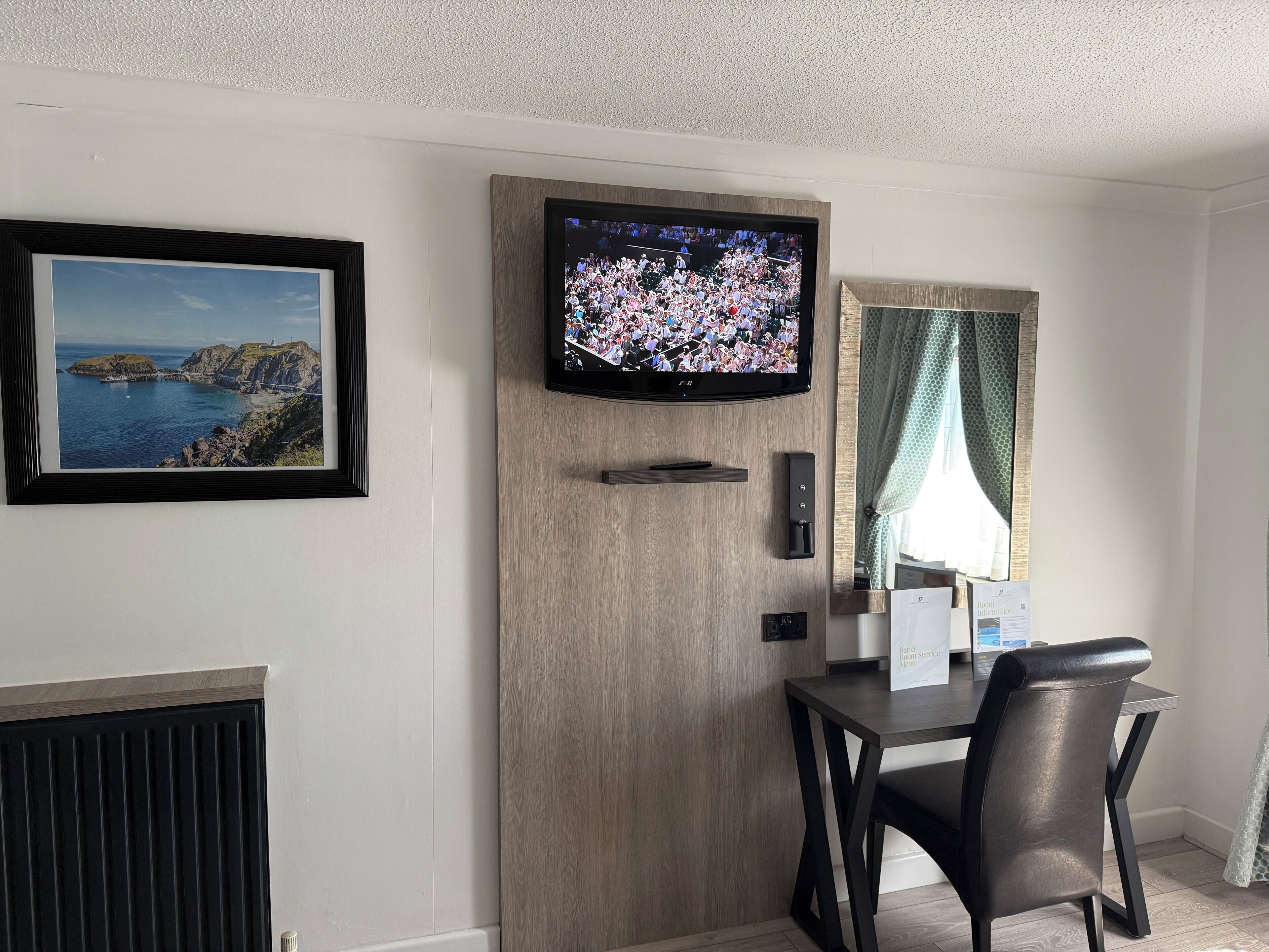 Habitació amb televisió penjada a un panell de fusta, un quadre amb un paisatge costaner, escriptori amb cadira i finestra amb cortines.