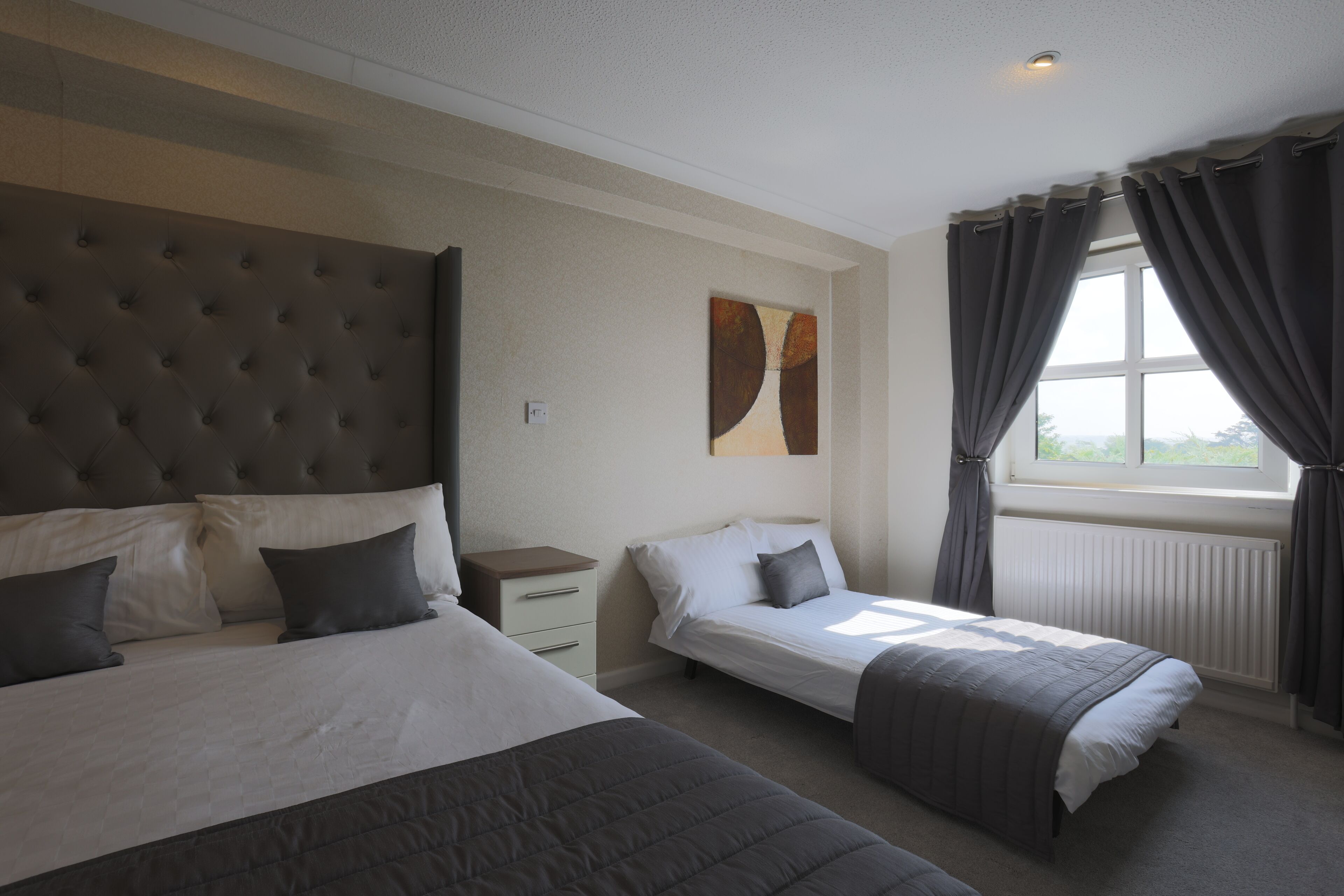 A bedroom with two beds, a nightstand, a window with curtains, and a wall-mounted artwork.