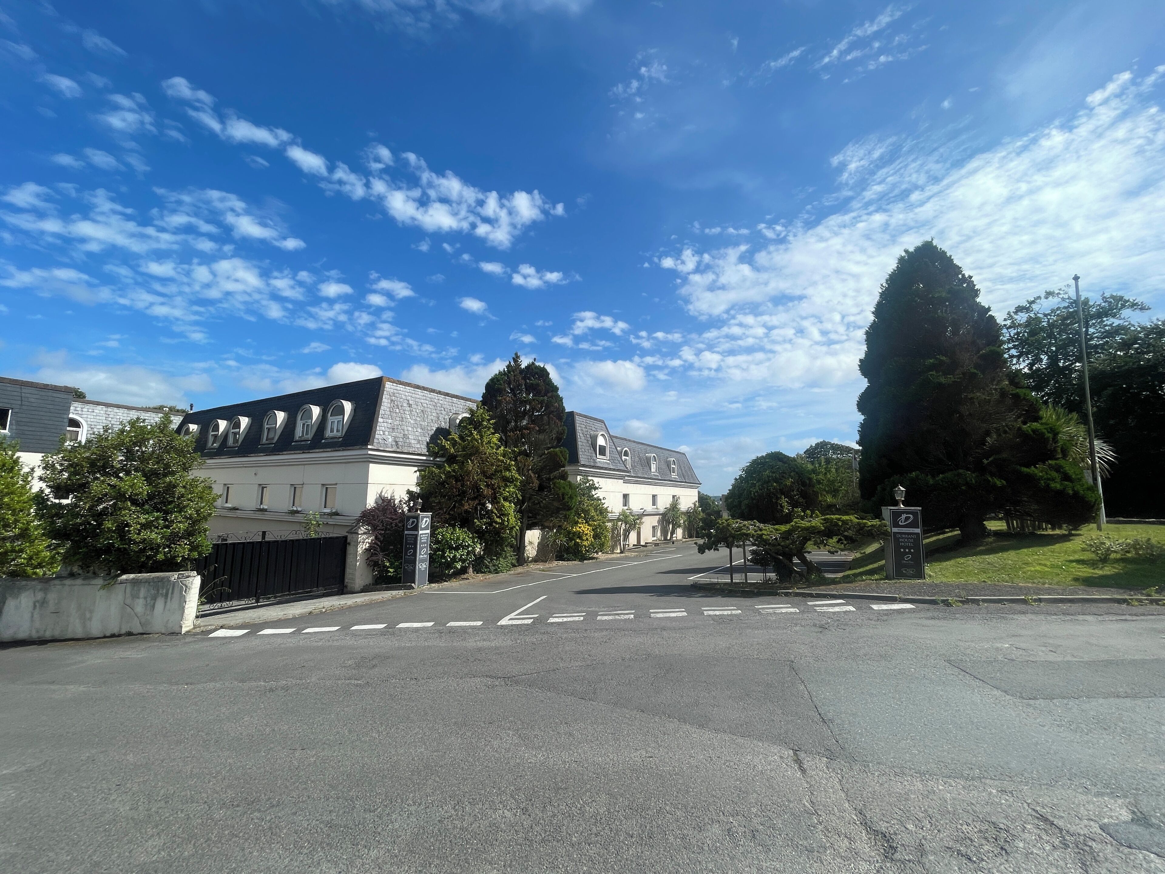 Straßenansicht eines Gebäudes mit symmetrischer Fassade, zahlreichen Fenstern und einem zentralen Eingang. Im Hintergrund sind Bäume und ein klarer Himmel zu sehen.