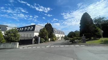 Straßenansicht eines Gebäudes mit symmetrischer Fassade, zahlreichen Fenstern und einem zentralen Eingang. Im Hintergrund sind Bäume und ein klarer Himmel zu sehen.