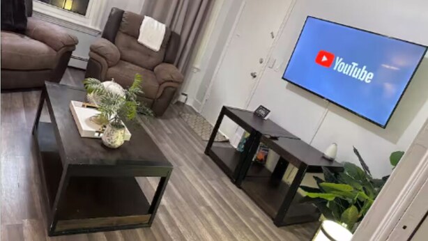 Un salon avec un téléviseur à écran plat fixé au mur, un canapé en cuir marron, une table basse agrémentée d’une plante et une petite table d’appoint.