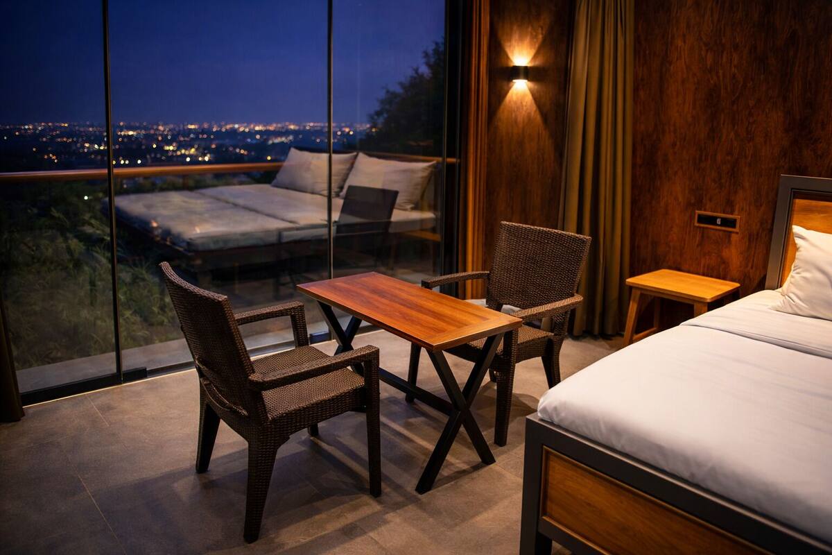 A bedroom with a bed, a table, two chairs, and a view of the city at night.