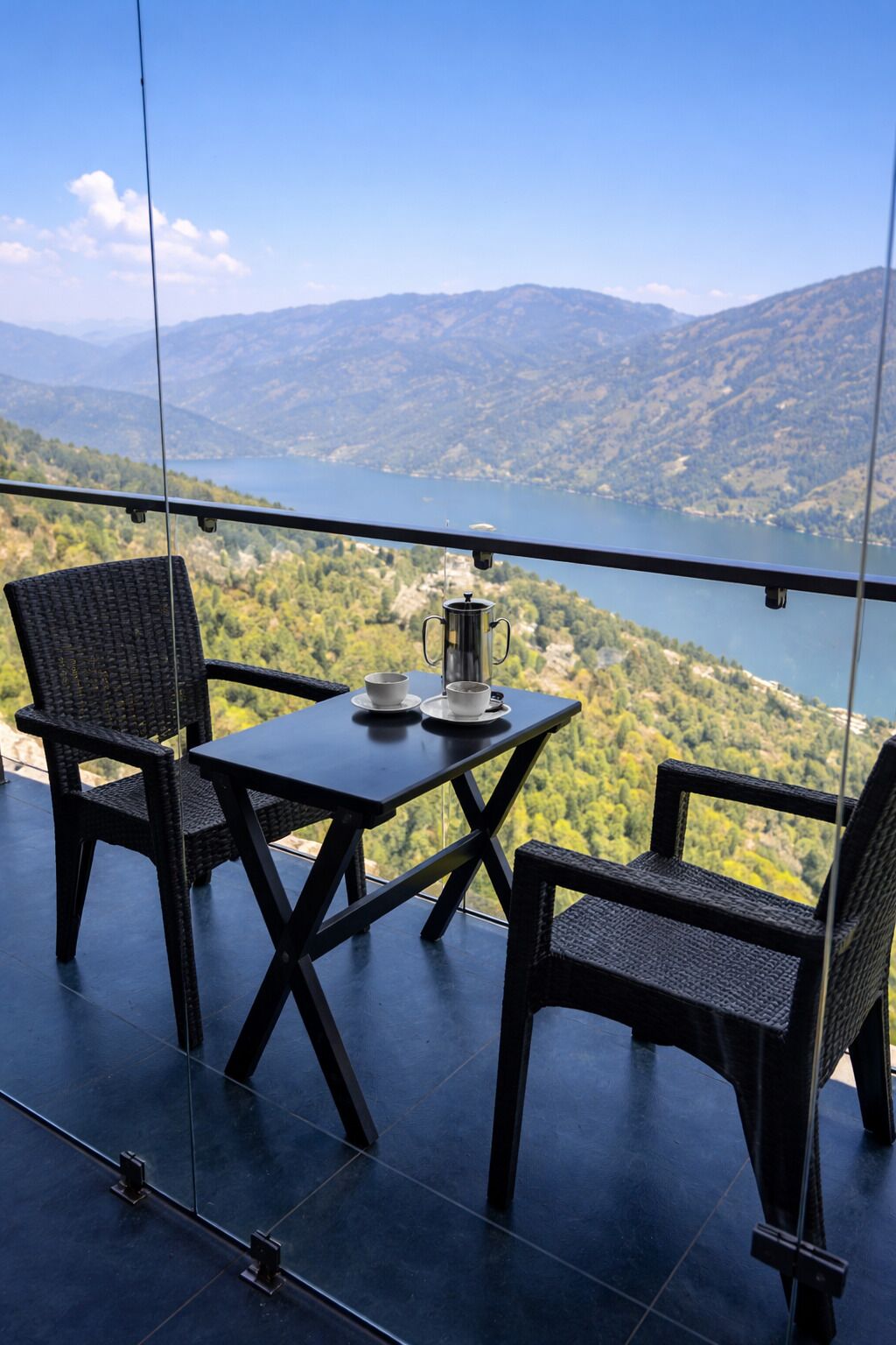 A balcony with a table and chairs, a metal coffee pot, and a view of a lake and mountains.