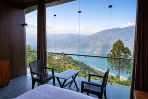 A balcony with a view of a lake and mountains, furnished with a bed, two chairs, and a table. - Ekaiva Resort, New Tehri : A Boutique Lake View Hotel (Tehri)