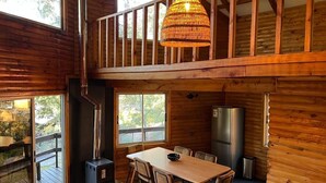A wooden cabin interior with a dining table, chairs, a hanging light fixture, a wood-burning stove, and a refrigerator.