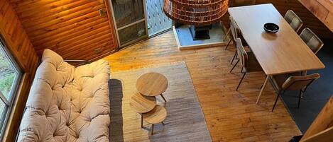 A cozy cabin interior with a cushioned sofa, a dining table with chairs, a woven lamp, and a view through a glass door.