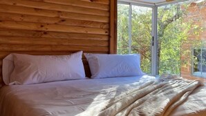 A bed with white linens and a beige blanket, wooden paneling on the wall, and a large window with a view of trees.