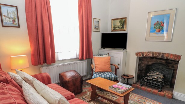 A cozy living room with a sofa, armchair, and a small table with a magazine. There is a fireplace, a television, and a window with curtains. - The Townhouse (Salisbury)