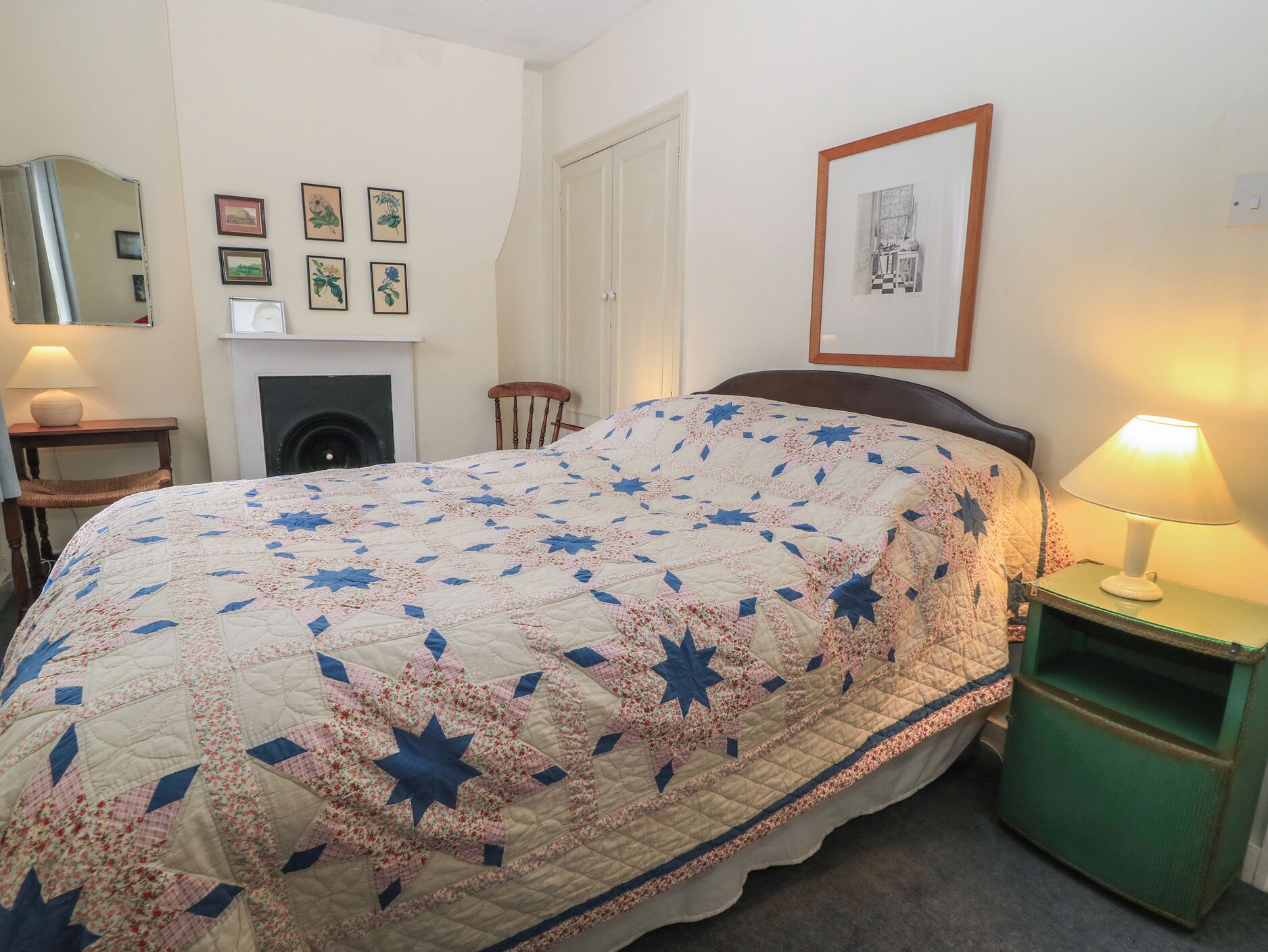 A bedroom with a bed, a nightstand, a lamp, a chair, a fireplace, and framed pictures on the wall.