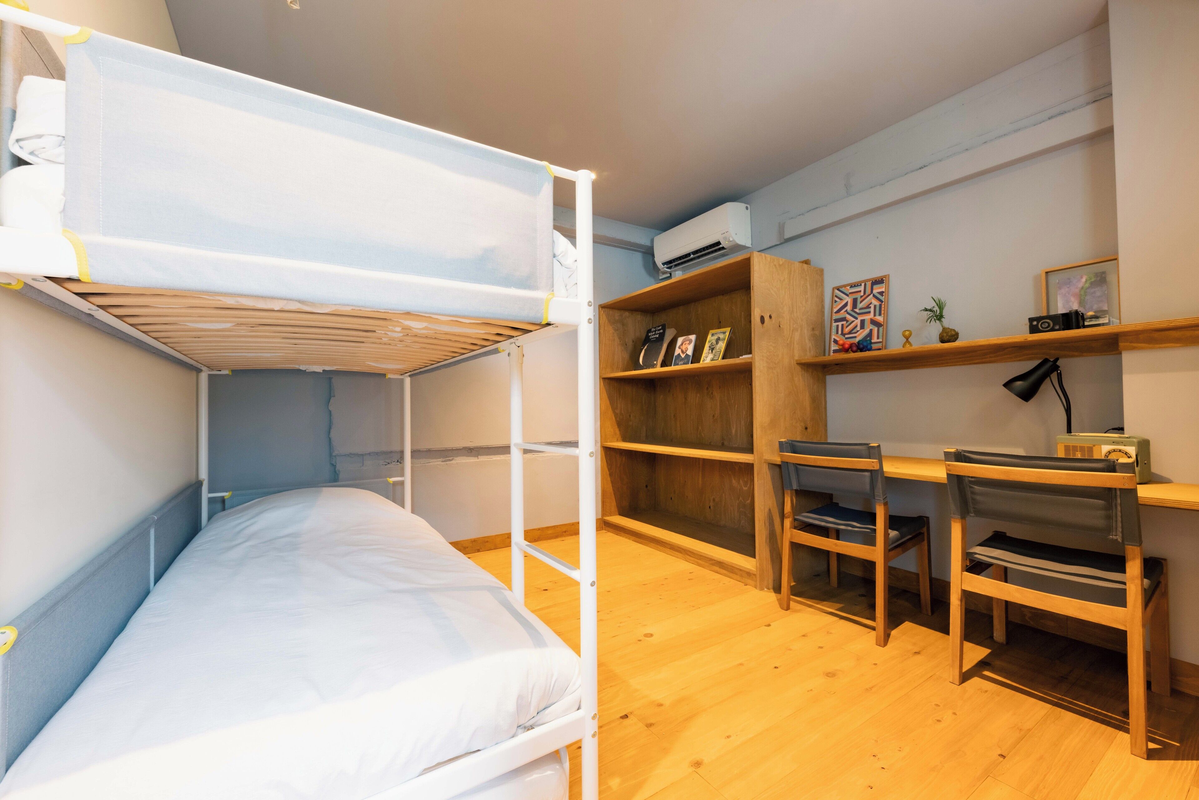 Habitación con literas, escritorio, sillas y estanterías de madera con objetos personales, y un equipo de aire acondicionado en la pared.