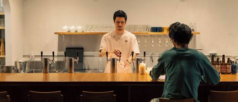 A bar area with a bartender serving a customer. There are bar stools, a counter, and shelves with glasses and bottles in the background.