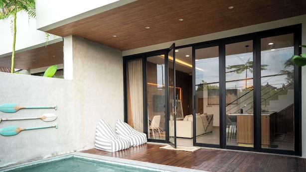 Un área de piscina moderna al aire libre con terraza de madera, zona de estar con cojines a rayas y vista del interior a través de puertas de cristal.