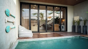 Un área de piscina moderna al aire libre con un deck de madera, una silla de descanso a rayas y vista a un interior residencial a través de amplios ventanales.
