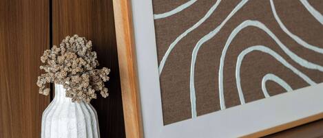 A framed artwork with abstract lines on a brown background, a white vase with dried flowers, and a wooden shelf against a wooden wall.